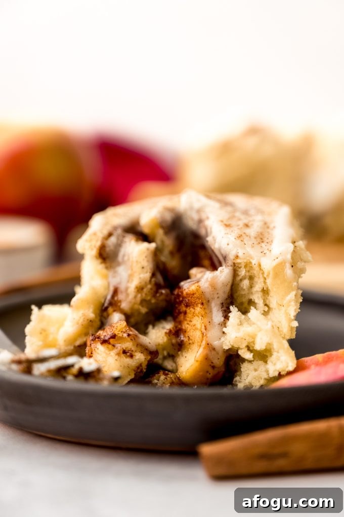 An apple cinnamon roll on a plate with a fork and apple slices and cinnamon sticks in the foreground.