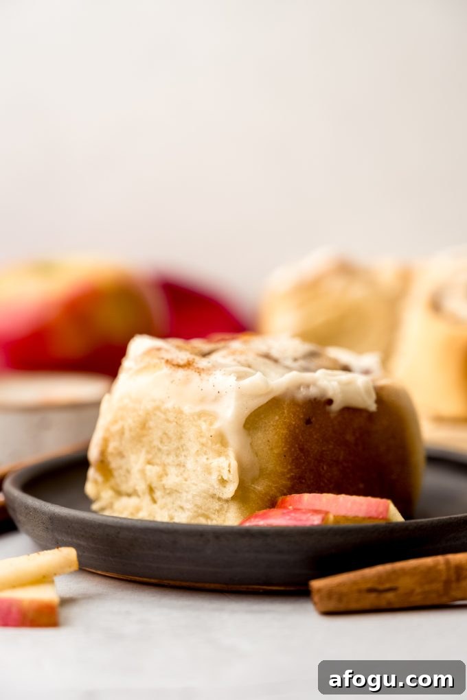 An apple cinnamon roll on a plate with apple slices and cinnamon sticks in the foreground.