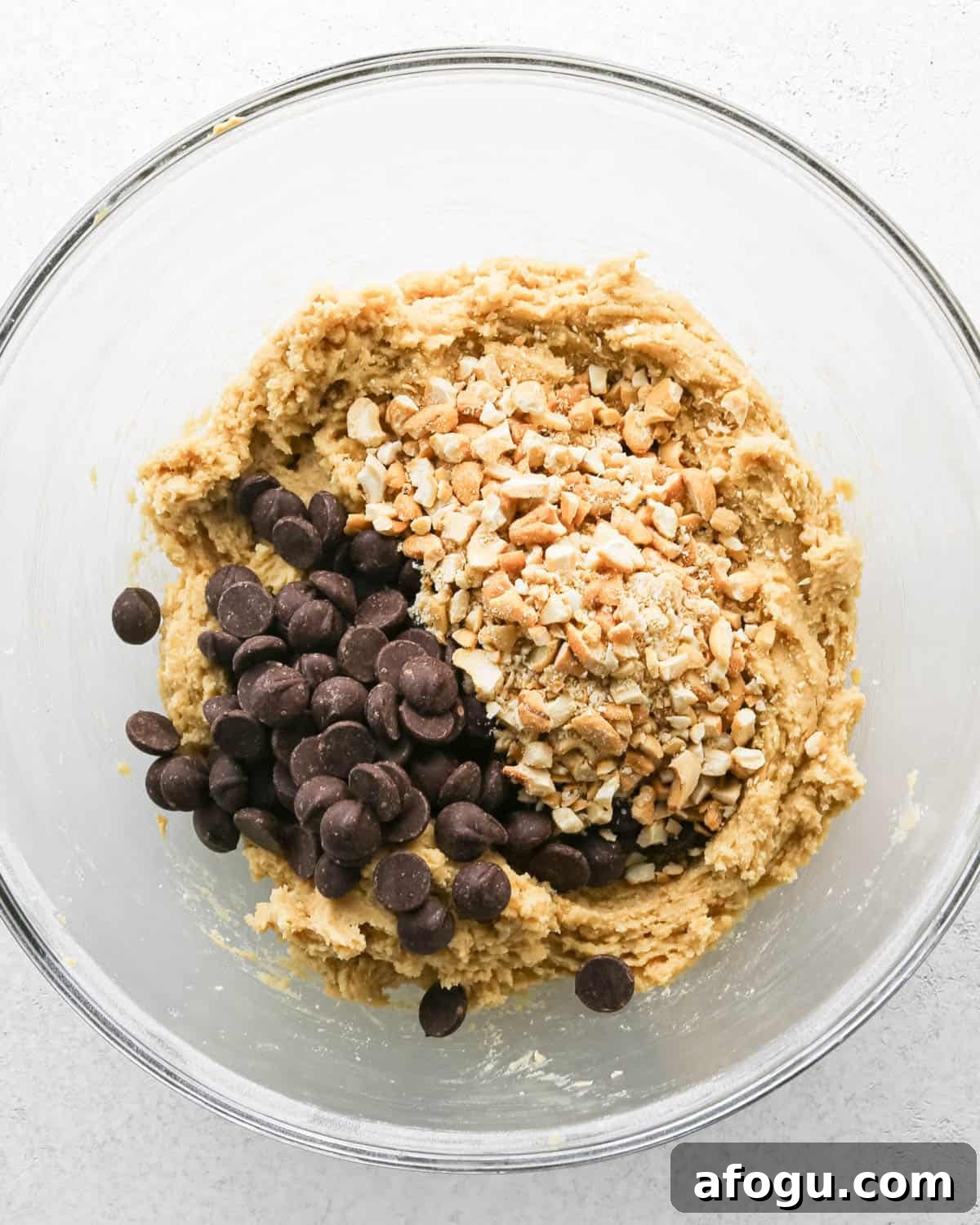 Chocolate Chip Cashew Cookies Recipe — Crunchy Homemade Treats 13 Making chocolate chip cashew cookies: overhead photo of chocolate chips and cashews added to the large clear mixing bowl with the cookie dough.