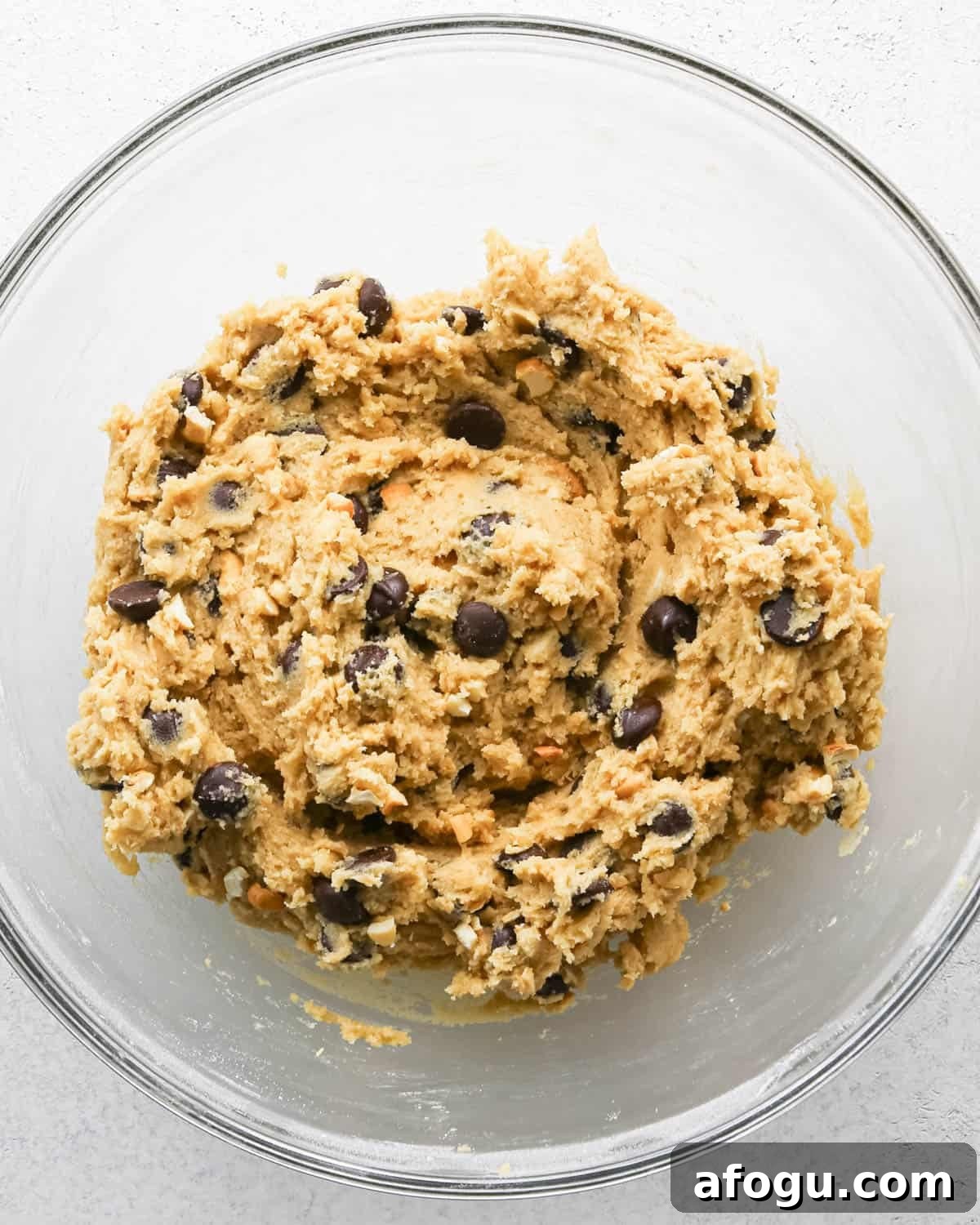 Chocolate Chip Cashew Cookies Recipe — Crunchy Homemade Treats 14 Making chocolate chip cashew cookies: overhead photo of chocolate chips and cashews added to the large clear mixing bowl with the cookie dough and mixed together.
