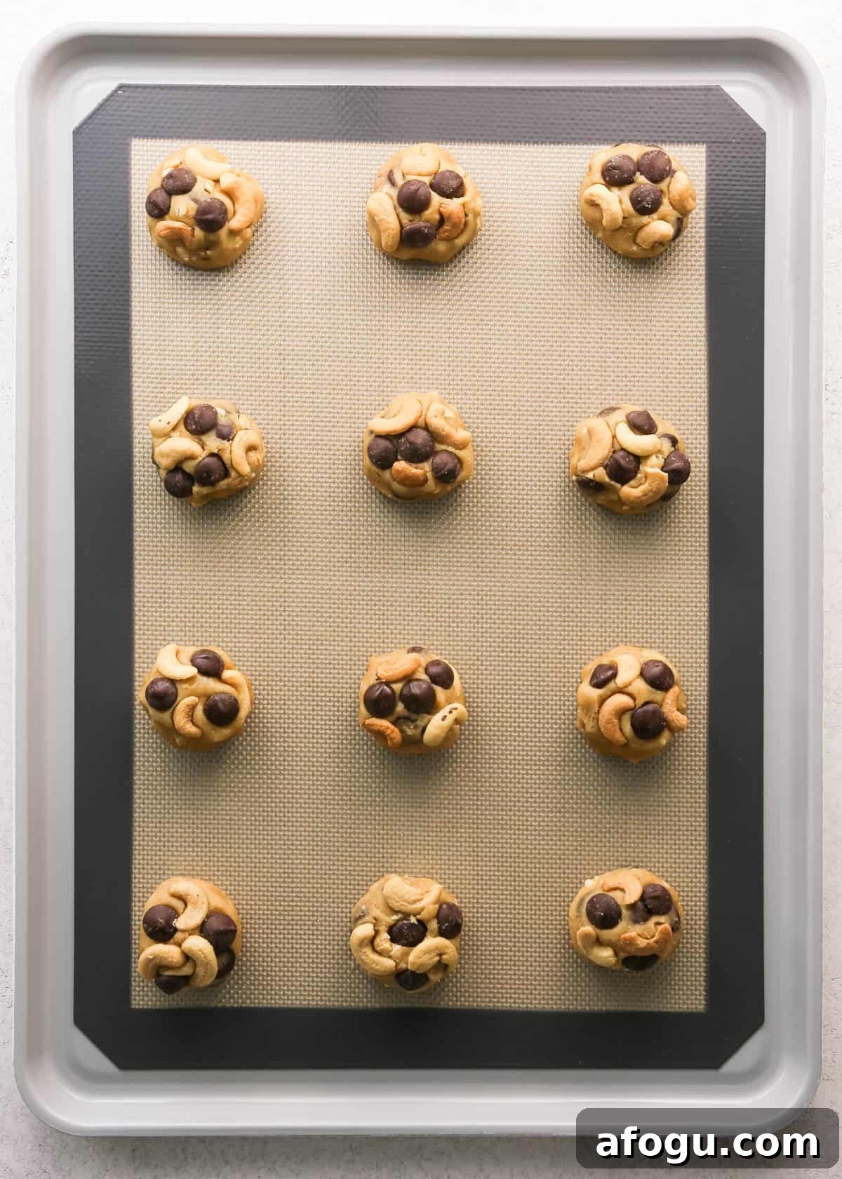 Chocolate Chip Cashew Cookies Recipe — Crunchy Homemade Treats 18 Making chocolate chip cashew cookies: overhead photo of 12 balls of cookie dough scooped, rolled, and placed onto a baking sheet on a countertop. Whole cashews have been added to the top of each cookie.