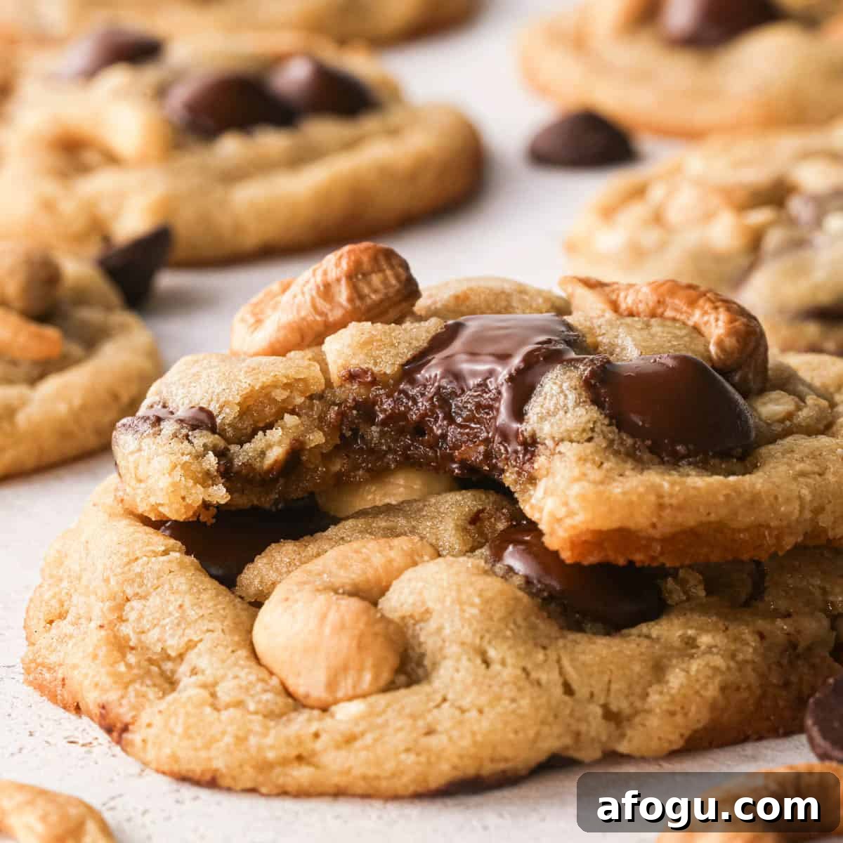 Chocolate Chip Cashew Cookies Recipe — Crunchy Homemade Treats 22 Up-close photo of a surface full of chocolate chip cashew cookies. A bite is taken out of one of the cookies to show the melty chocolate, the crispy outside, and the chewy, perfectly gooey inside of the cookie. Extra chocolate chips and cashews surround the cookies on a white countertop.