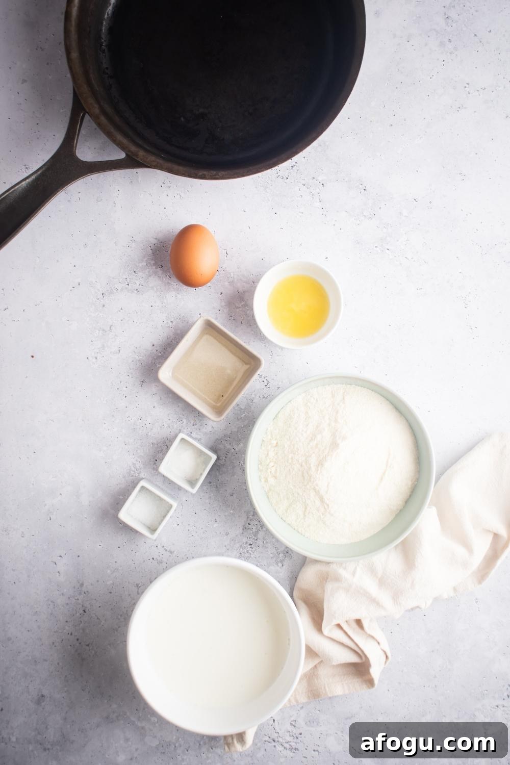 An egg, a bowl of melted butter, a bowl of flour, and a bowl of buttermilk all on a white counter.