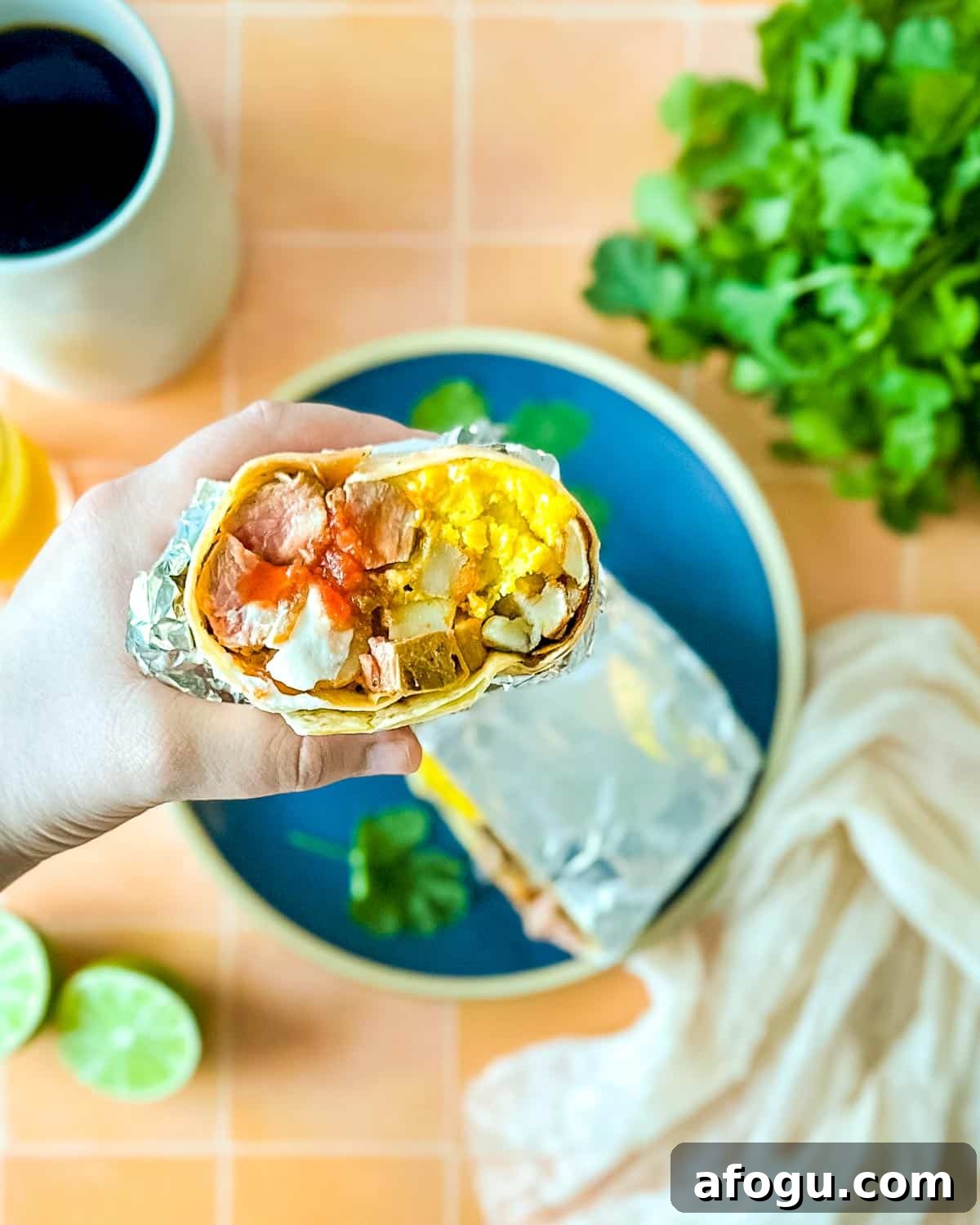 Half of a carne asada breakfast burrito is held over a blue plate that's surrounded by limes, coffee, and orange juice.