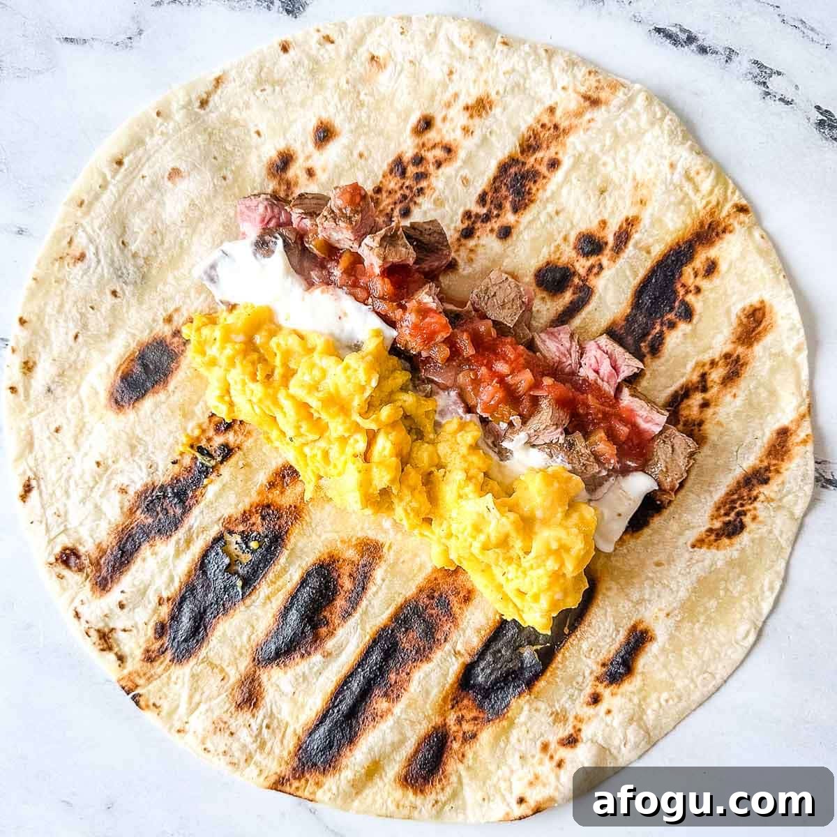 Eggs, carne asada, sour cream, and salsa sit on a grilled flour tortilla.