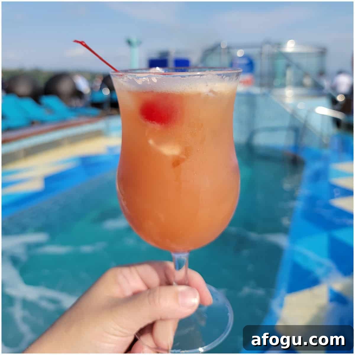 pink orange cocktail in a hurricane glass with a cherry in front of a pool