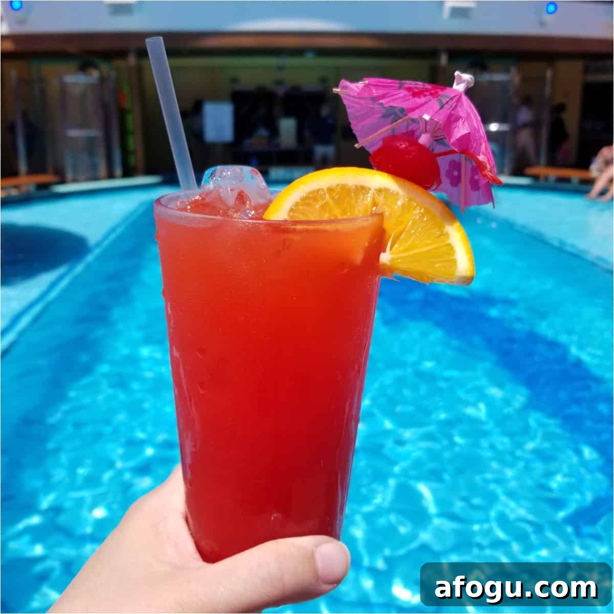 hand holding a red cocktail with orange wheel and pink umbrella by the pool