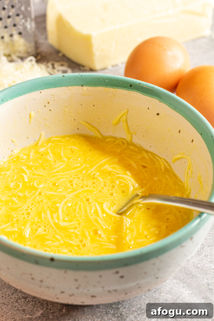 A ceramic ivory bowl with black specks and an aquamarine rim sits on a gray marbled counter. Inside the bowl is a mixture of eggs and cheese that has been stirred with a fork. Behind the bowl are two brown eggs, and a block of mozzarella cheese that has been partially grated.