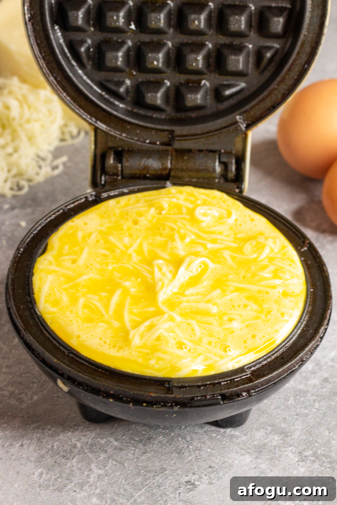 a mini waffle iron is open and filled with a mixture of eggs and cheese. In the background you can see 2 brown eggs and a pile of finely grated white cheese
