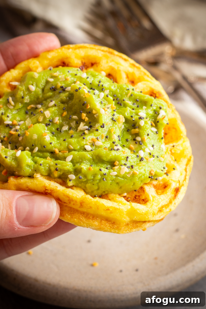 a hand holds a small round chaffle. It is topped with mashed avocado and sprinkled with everything bagel seasoning