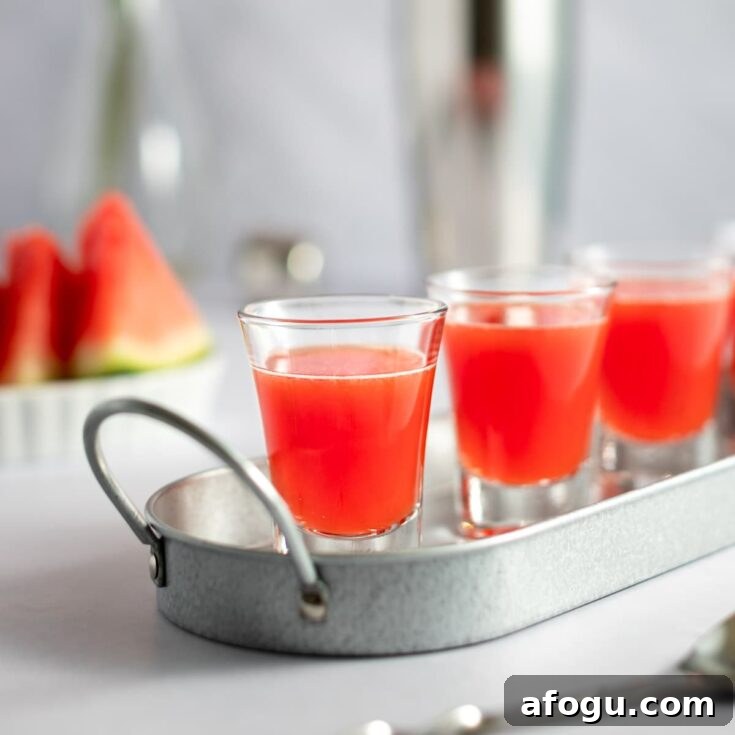Watermelon shooters lined up on a metal tray, with sliced watermelon and a cocktail shaker in the background.