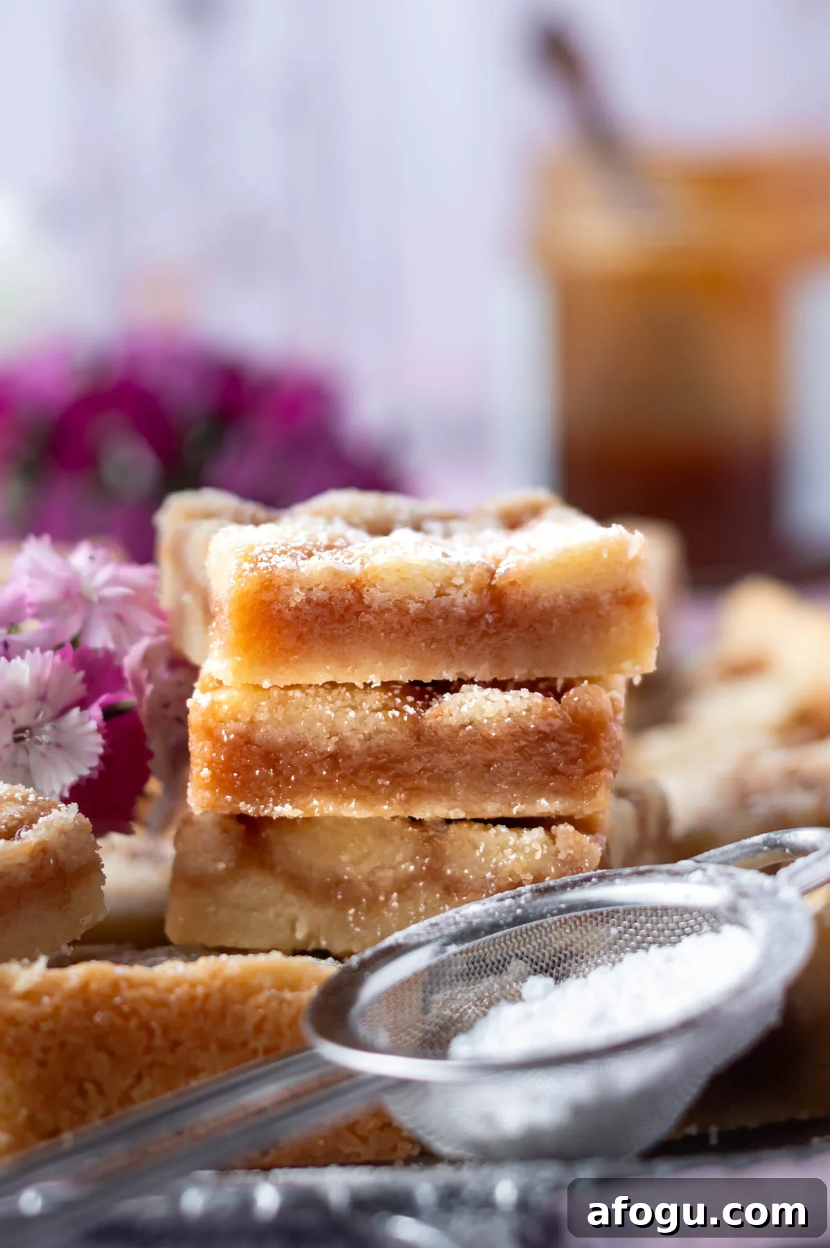 stack of cookie bars with a jar of caramel in the background and confectioners' sugar in a sifter