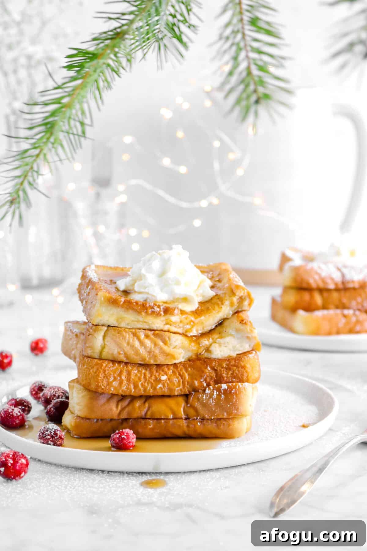 Eggnog French Toast Recipe: Festive Holiday Brunch Idea 2 french toast on a white plate with greenery, more toast behind, fairy lights, and cranberries