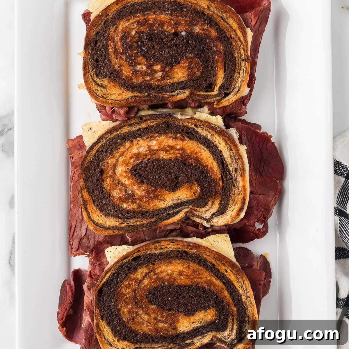Three Blackstone Reuben Sandwiches made with swirled rye bread are stacked vertically on a white rectangular plate, showcasing layers of corned beef, cheese, and other traditional fillings.