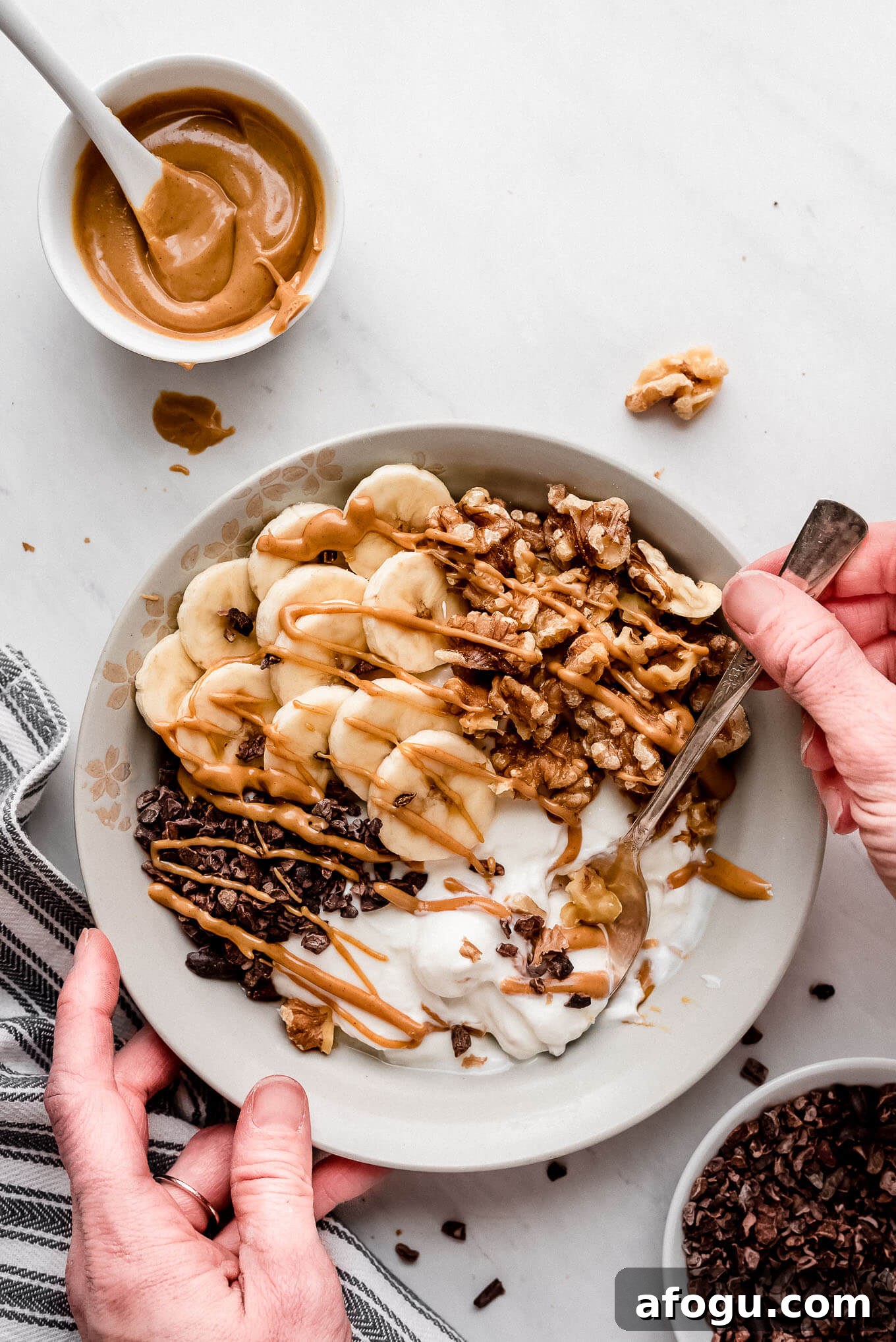 Spoonful of yogurt bowl. Hands holding a bowl and lifting a spoonful of yogurt from a Greek yogurt bowl topped with bananas, chocolate, and nuts.