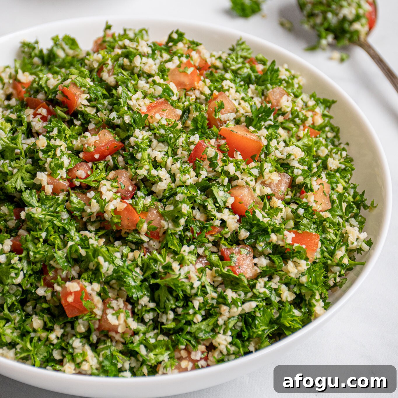 A large white bowl filled with tabbouleh salad with a spoonful out on the side with parsley leaves falling off.