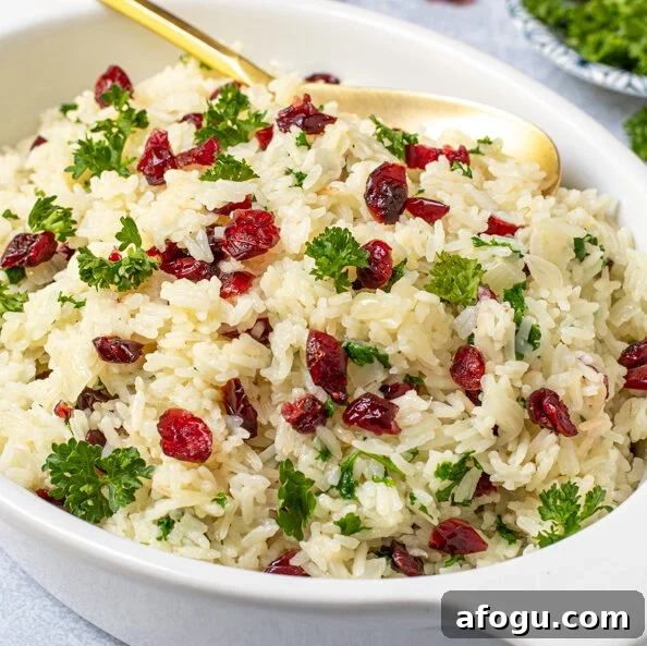 A white serving dish with a gold spoon in white rice pilaf with dried cranberries and fresh parsley.