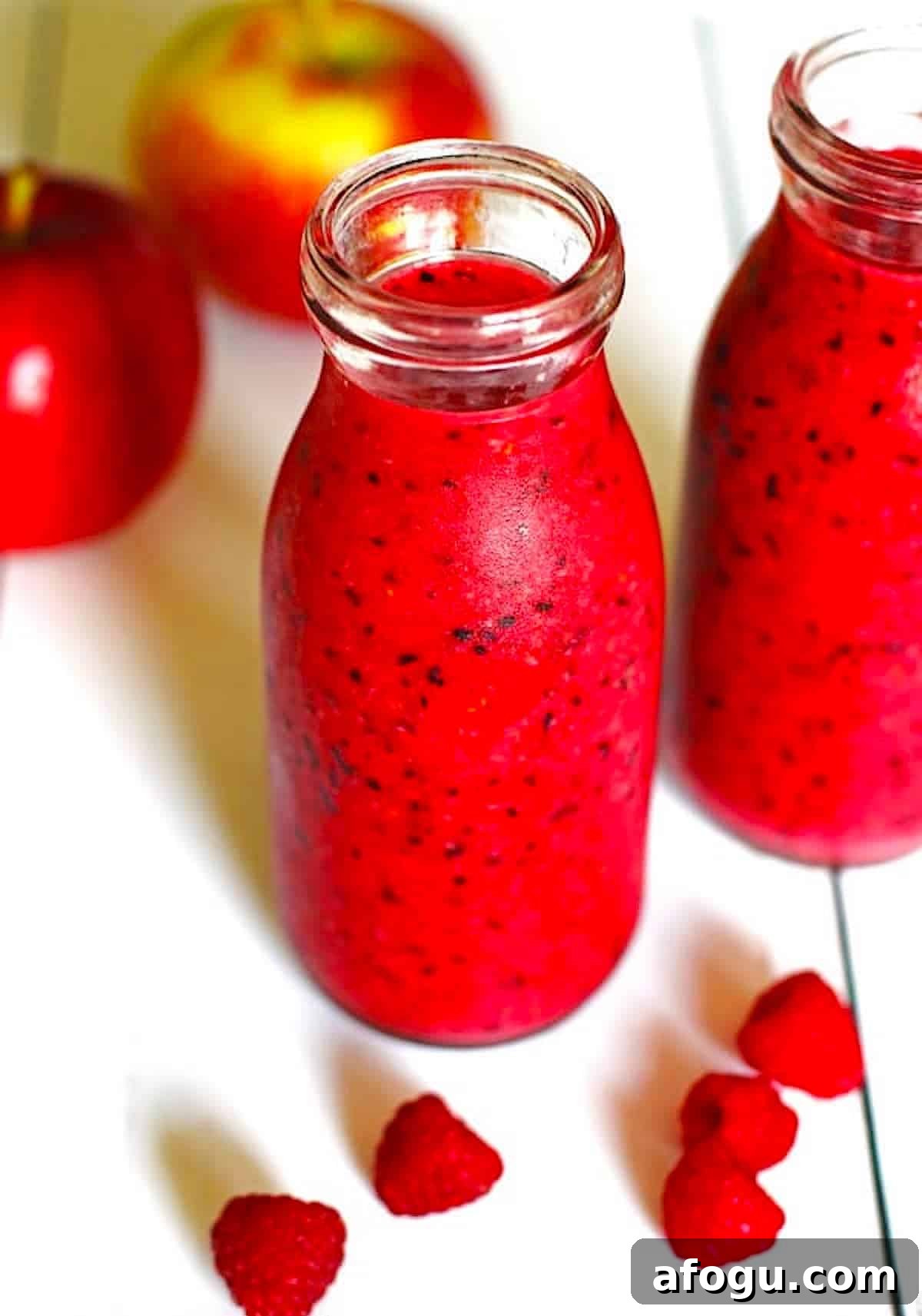Apple Smoothie in two clear glasses on a white table with apples and raspberries next to the smoothies.