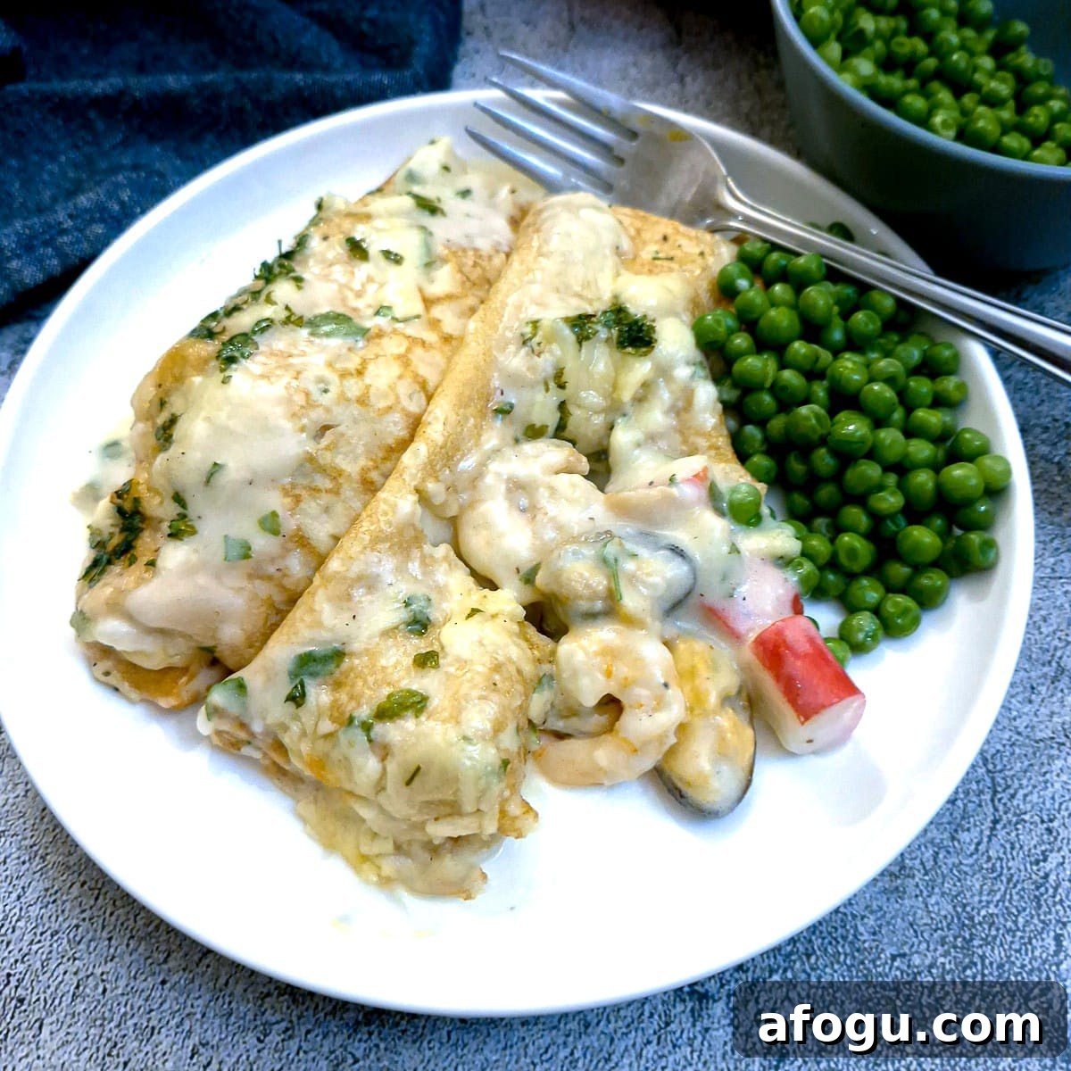 2 seafood stuffed pancakes with peas on a white plate.  One pancake has been cut in half to show the filling.