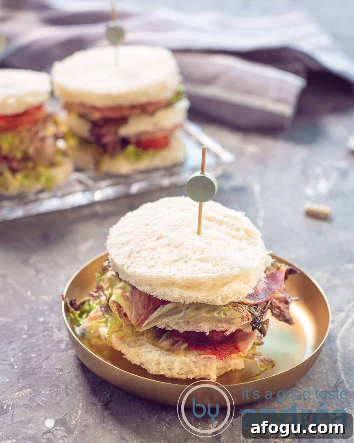 A photo in the height of a bowl of gold with a bacon lettuce and tomato sandwich. On a grey background More sandwiches in the back. 