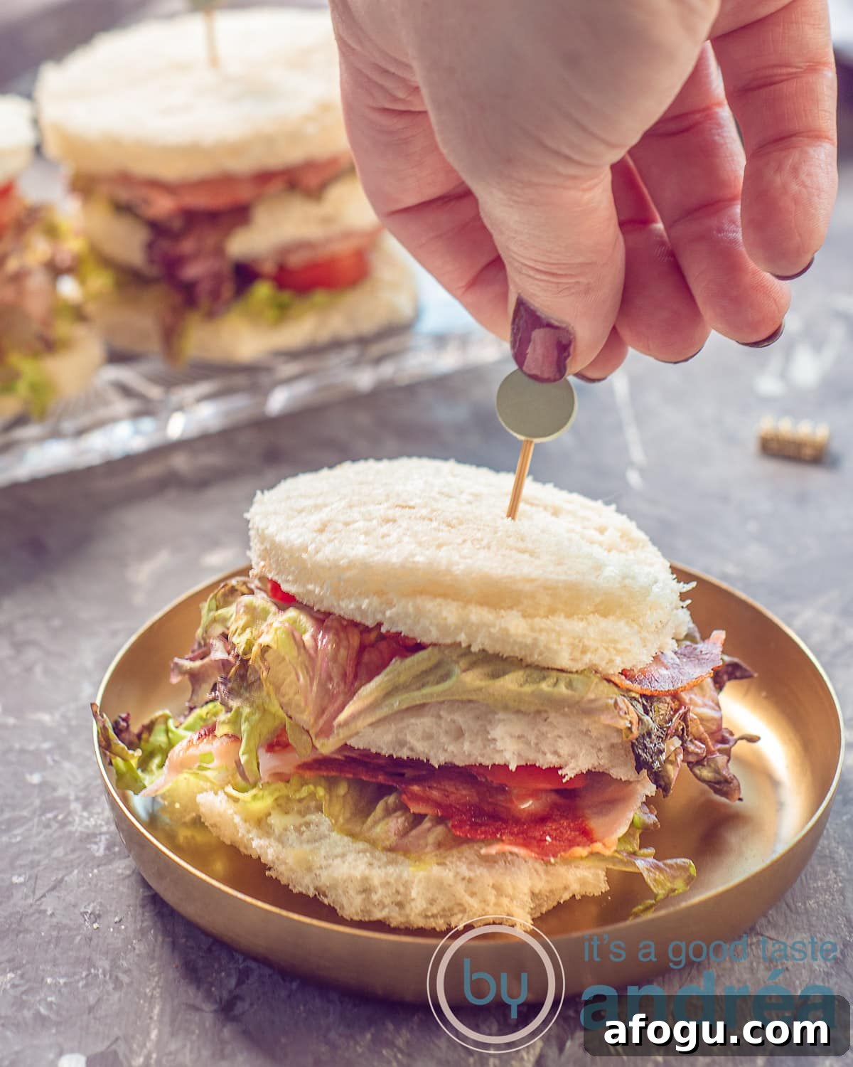 A photo in height with a gold plate with a BLT mini Sandwich on it. A hand sticks a tapas picker in it. More sandwiches in the background