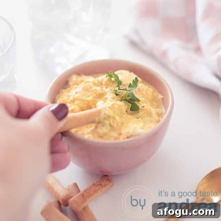 A square photo of a pink container filled with an egg salad without mayonnaise. A tapas stick scoops something out. A white background. Top right a pink white tea towel