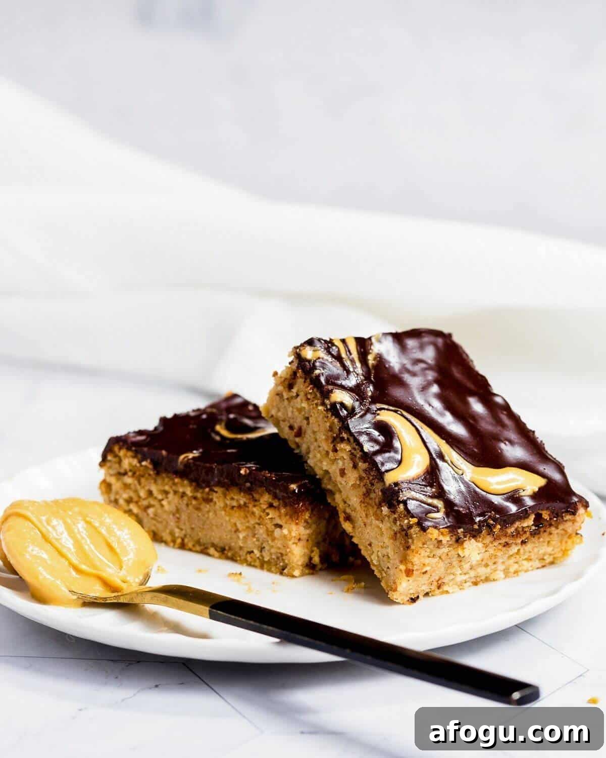 Two slices of peanut butter cake on a white plate. There is a spoon with peanut butter next to the cake.