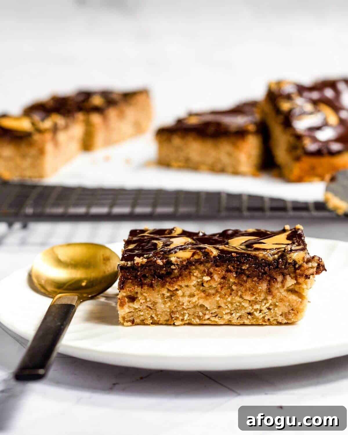 A slice of peanut butter cake on a white plate, there is a spoon next to the cake on the plate. In the background the rest of the cake is on a wire rack.