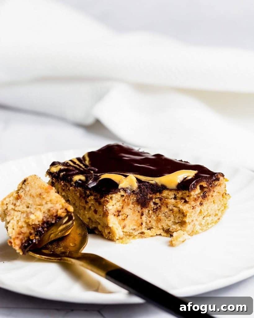 A slice of peanut butter cake with chocolate ganache on a white plate. There is a spoon on the plate with a mouthful of cake on it.