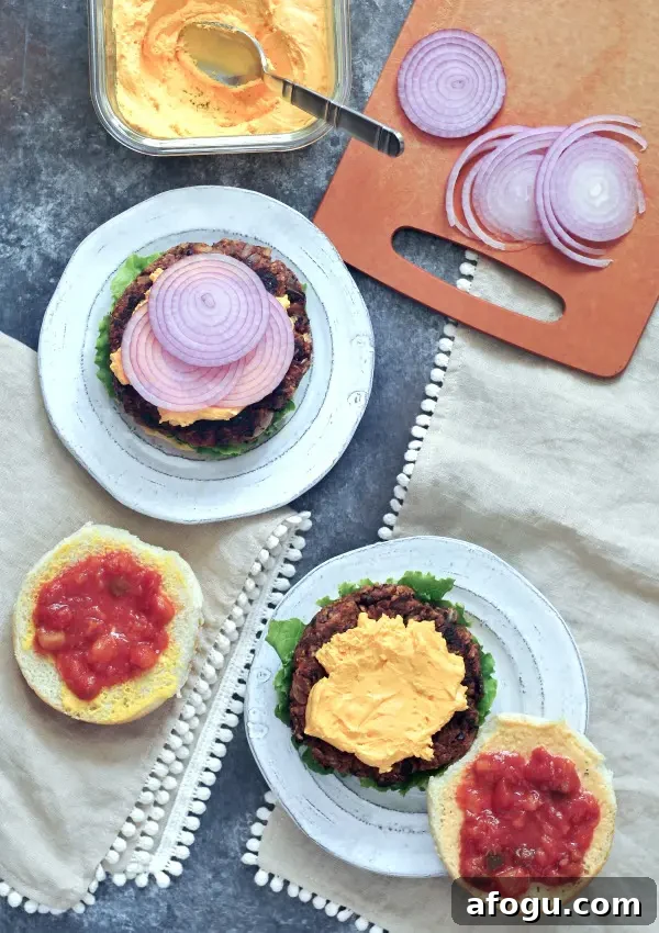 Overhead view of southwest black bean burgers sitting on plates on a picnic table. Burgers are dressed with curly lettuce, cheese sauce, salsa, purple onion slices and sesame seed buns.
