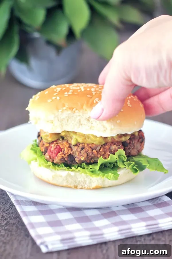 A hand is picking up a southwest black bean burger dressed with lettuce, cheese sauce, green chiles and a sesame seed bun.