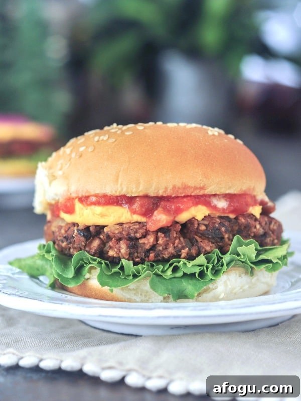 A salsa black bean burger sits on a plate on a picnic table. Burger is dressed with curly lettuce, cheese sauce, salsa, and a sesame seed bun.