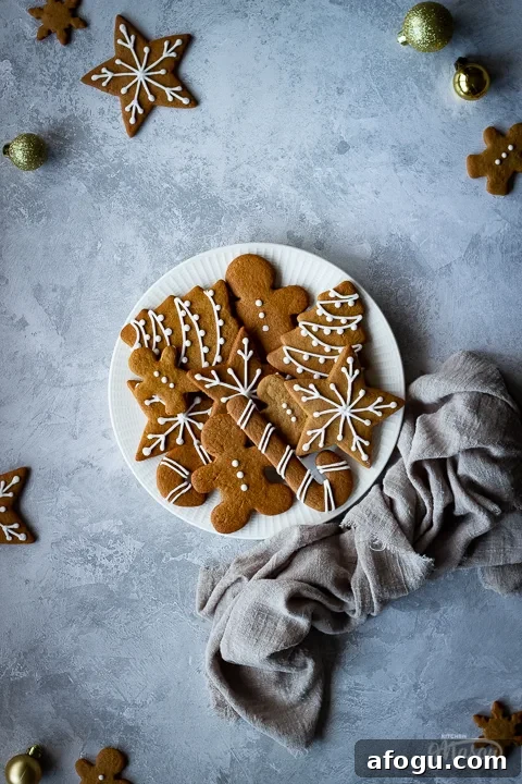 Classic Gingerbread Biscuit Recipe for Perfect Holiday Baking 6 A white plate filled with gingerbread biscuits with a light brown linen napkin scrunched up to the side. There are more biscuits and gold baubles scattered around and it's all set on a grey plaster effect backdrop.