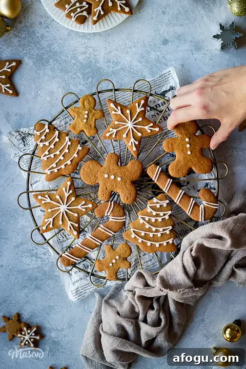 Classic Gingerbread Biscuit Recipe for Perfect Holiday Baking 7 A hand reaching into a cooling rack topped with iced gingerbread biscuits set over a scrunched up piece of white baking paper. In the background there is a light brown linen napkin, more iced gingerbread biscuits, some gold baubles, cookie cutters and a white plate with more biscuits on it. All set on a grey plaster effect backdrop.