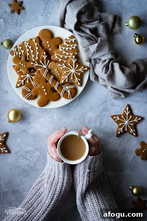 Classic Gingerbread Biscuit Recipe for Perfect Holiday Baking 2 Hands holding a cup of tea next to a white plate filled with decorated gingerbread biscuits with a light brown linen napkin scrunched up to the side. There's also more biscuits and gold baubles scattered in the background. All set over a grey plaster effect backdrop.