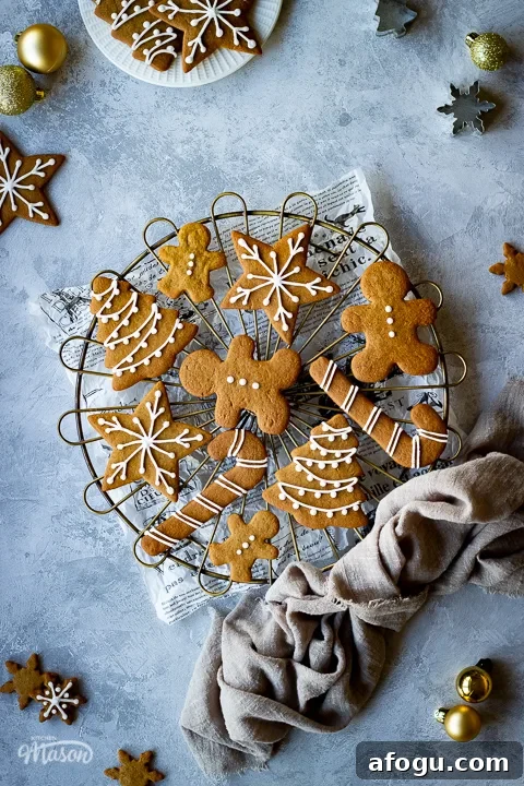 Classic Gingerbread Biscuit Recipe for Perfect Holiday Baking 3 A cooling rack topped with iced gingerbread biscuits set over a scrunched up piece of white baking paper. In the background there is a light brown linen napkin, more iced gingerbread biscuits, some gold baubles, cookie cutters and a white plate with more biscuits on it. All set on a grey plaster effect backdrop.