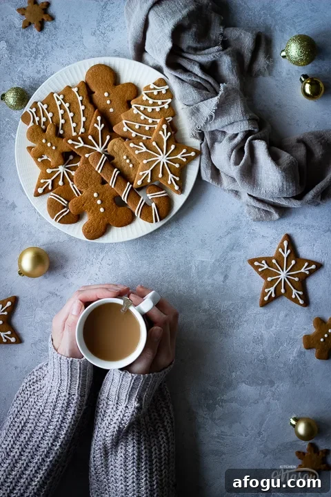 Classic Gingerbread Biscuit Recipe for Perfect Holiday Baking 18 Hands holding a cup of tea off to the side, next to a white plate filled with decorated gingerbread biscuits with a light brown linen napkin scrunched up to the side. There's also more biscuits and gold baubles scattered in the background. All set over a grey plaster effect backdrop.