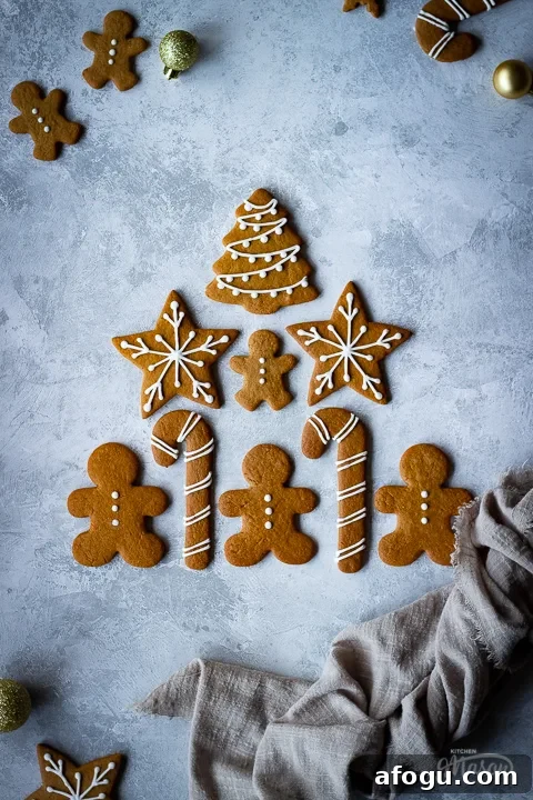 Classic Gingerbread Biscuit Recipe for Perfect Holiday Baking 19 9 decorated gingerbread biscuits laid on a grey plaster effect backdrop in a triangle shape with a light brown linen napkin scrunched up to the side. There are more biscuits and gold baubles in the background.