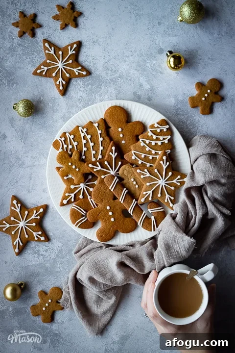 Classic Gingerbread Biscuit Recipe for Perfect Holiday Baking 20 A white plate filled with gingerbread biscuits with a light brown linen napkin scrunched up to the side. There are more biscuits, gold baubles and a hand holding a cup of tea in the background and it's all set on a grey plaster effect backdrop.