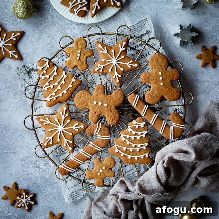 A cooling rack topped with iced gingerbread biscuits set over a scrunched up piece of white baking paper. In the background there is a light brown linen napkin, more iced gingerbread biscuits, some gold baubles, cookie cutters and a white plate with more biscuits on it. All set on a grey plaster effect backdrop.
