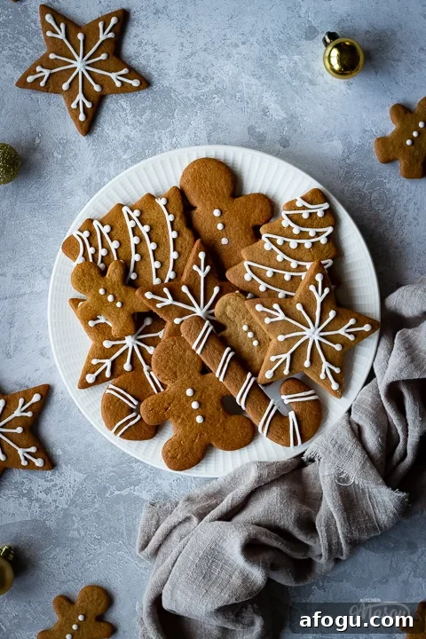 Classic Gingerbread Biscuit Recipe for Perfect Holiday Baking 4 A close up of a white plate filled with gingerbread biscuits with a light brown linen napkin scrunched up to the side. There are more biscuits and gold baubles scattered around and it's all set on a grey plaster effect backdrop.