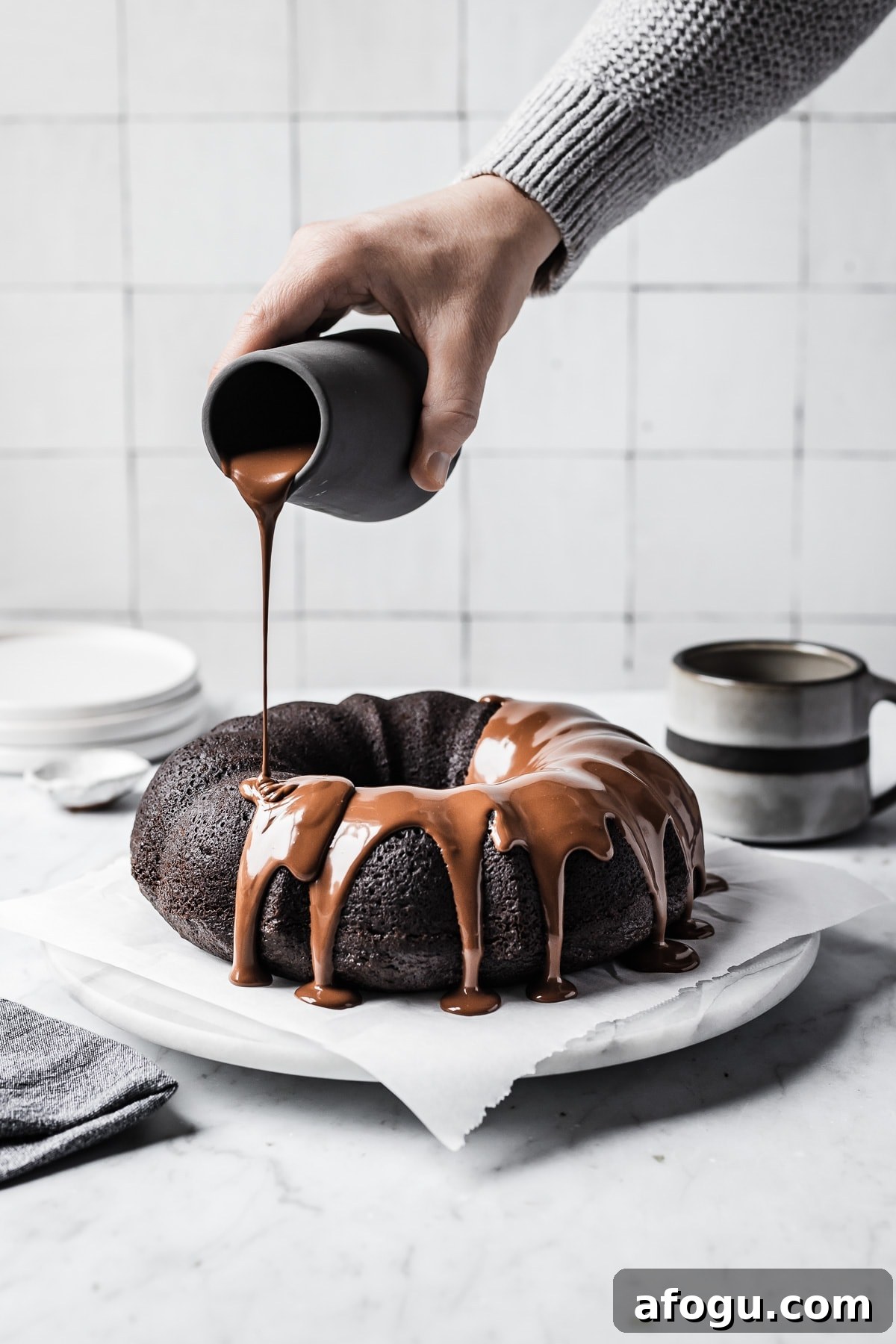 Hand pouring chocolate ganache glaze onto a chocolate olive oil bundt cake resting on a white marble platter