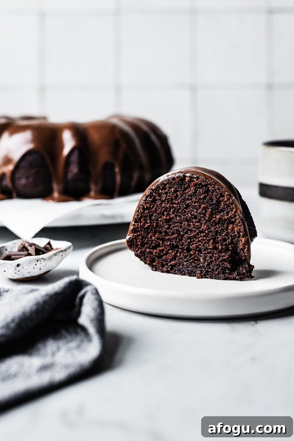 Slice of chocolate olive oil bundt cake on a white plate and marble counter with whole cake on platter in the background