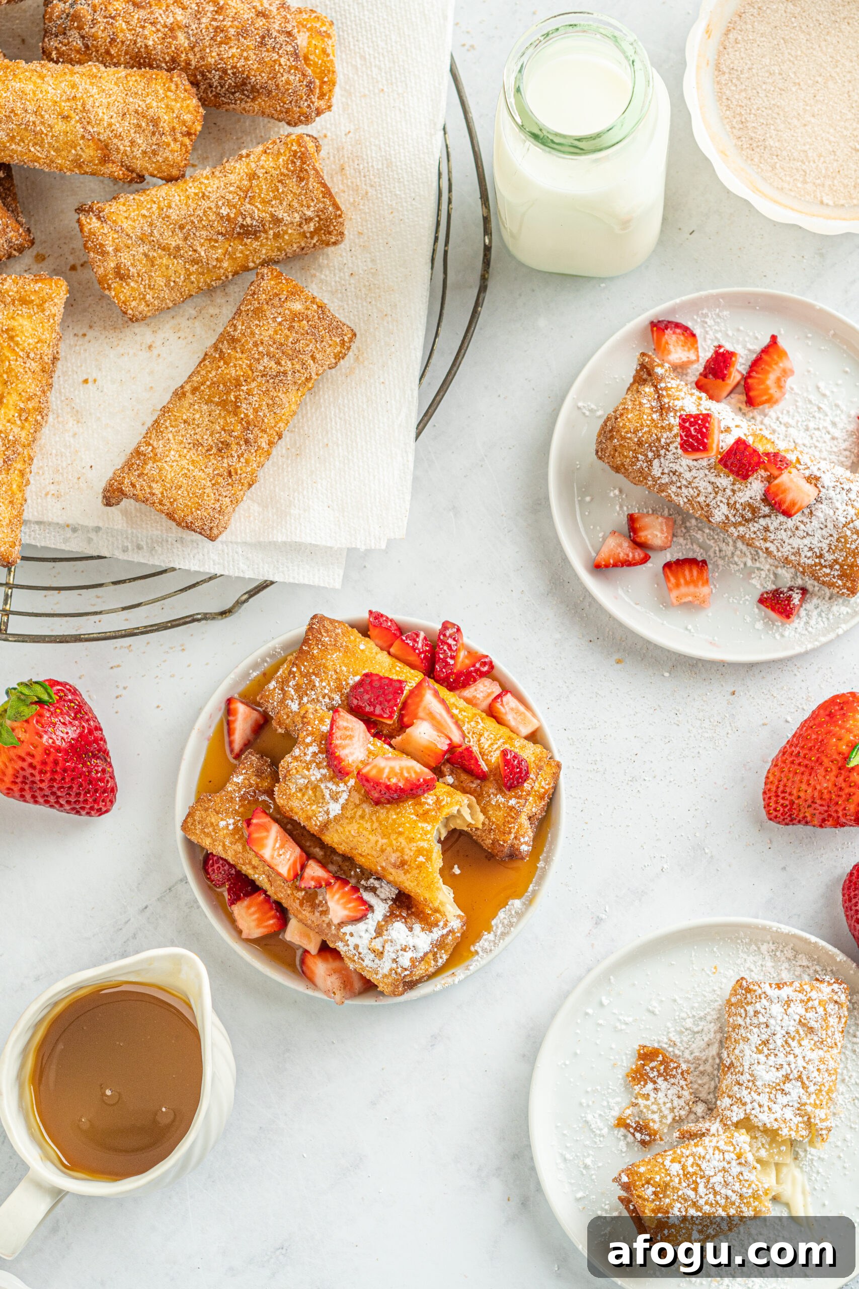 Overhead view of Cheesecake Egg Rolls stacked on a plate covered with powdered sugar, strawberries and caramel, with a bite removed from the top egg roll.