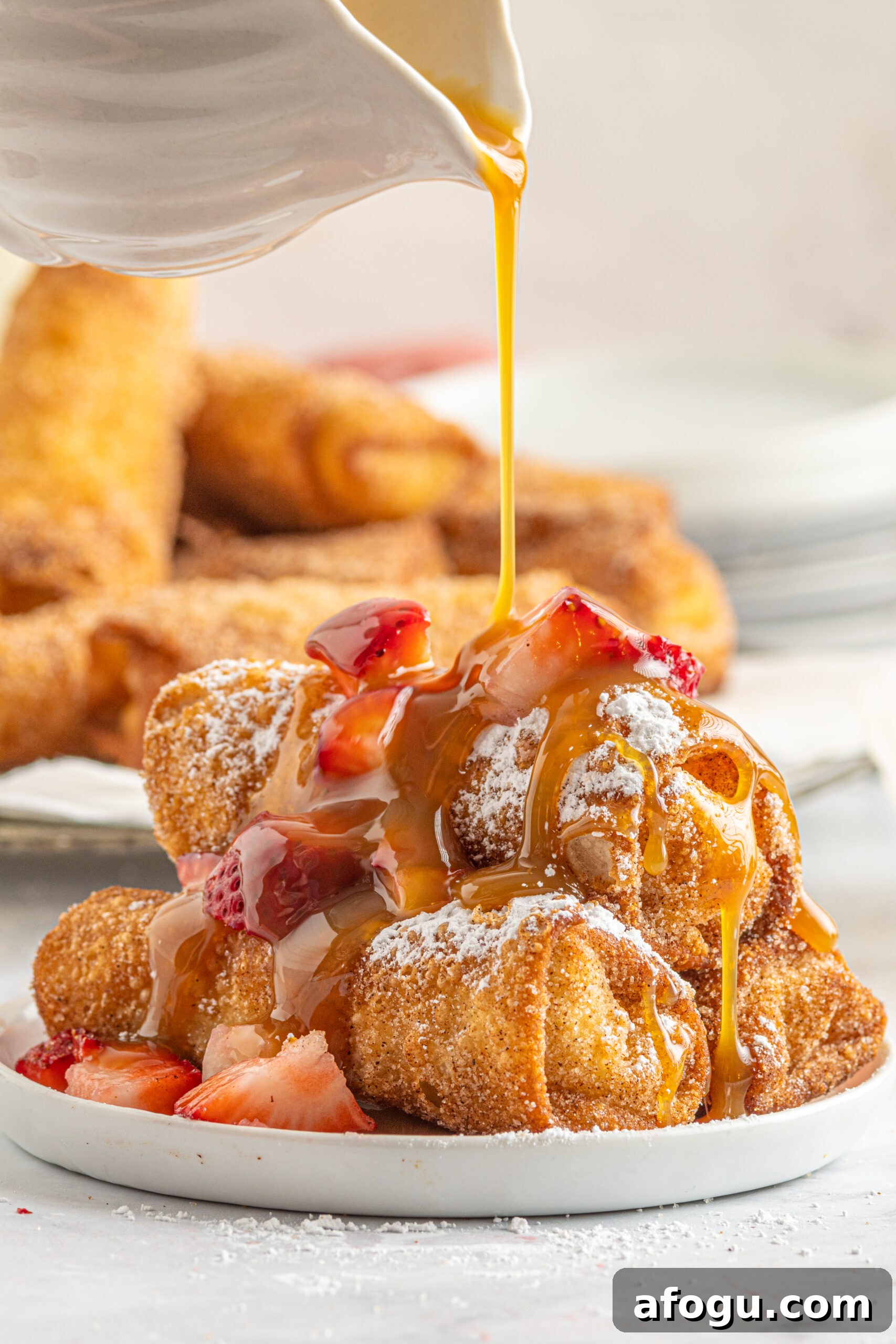 Front view of Cheesecake Egg Rolls stacked on a plate covered with powdered sugar, strawberries and caramel.