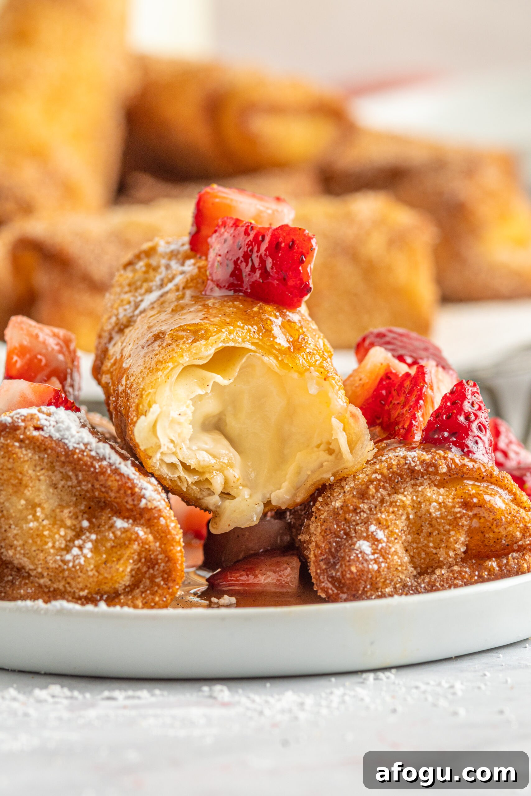 Front view of Cheesecake Egg Rolls stacked on a plate covered with powdered sugar, strawberries and caramel, with a bite removed from the top egg roll.