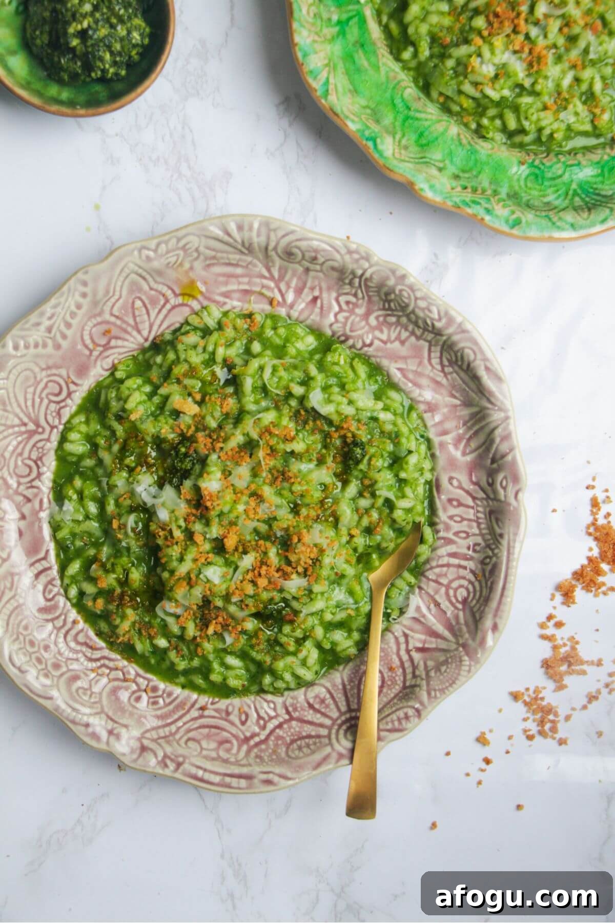 Wild garlic risotto topped with crispy breadcrumbs on a small pink plate, with another plate of risotto in the background.