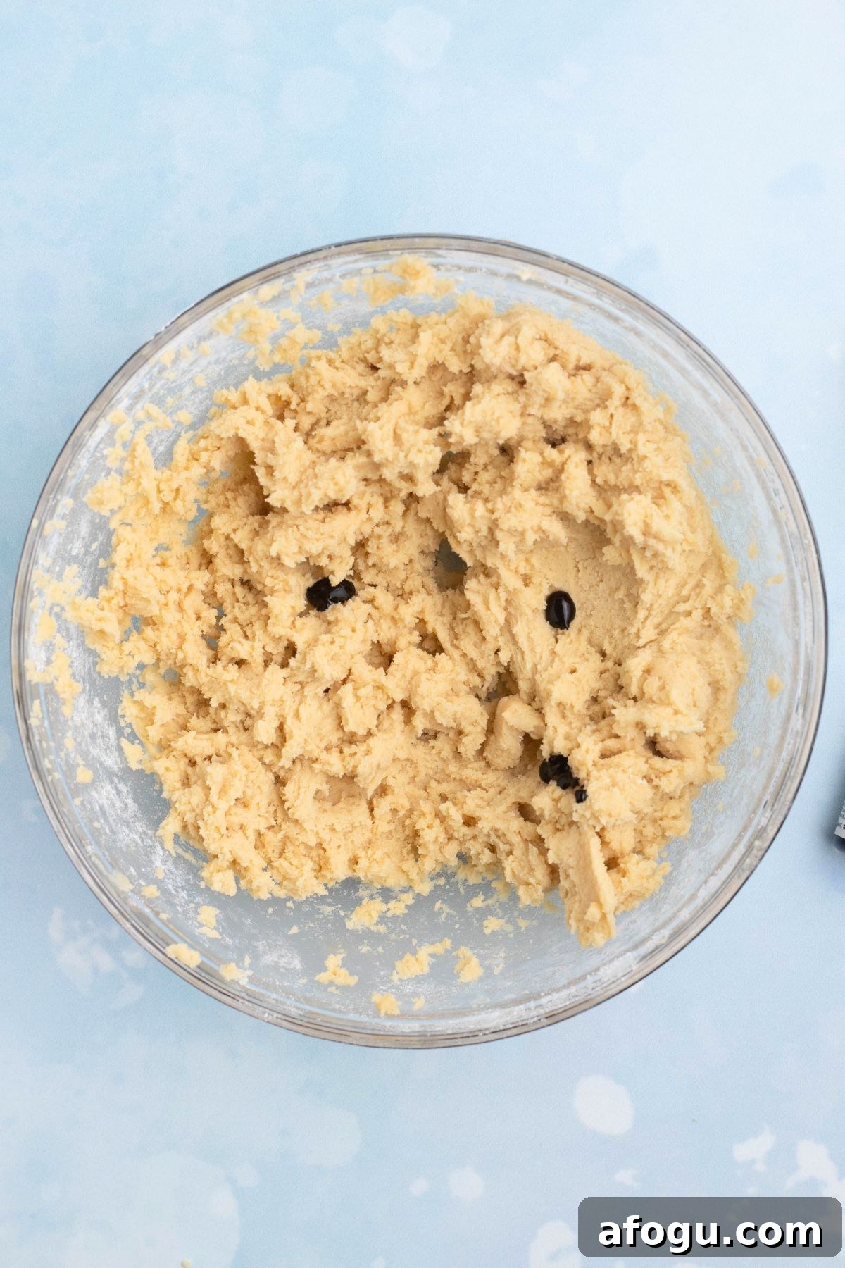 Cookie dough being mixed with royal blue and violet food colors in a clear glass mixing bowl.