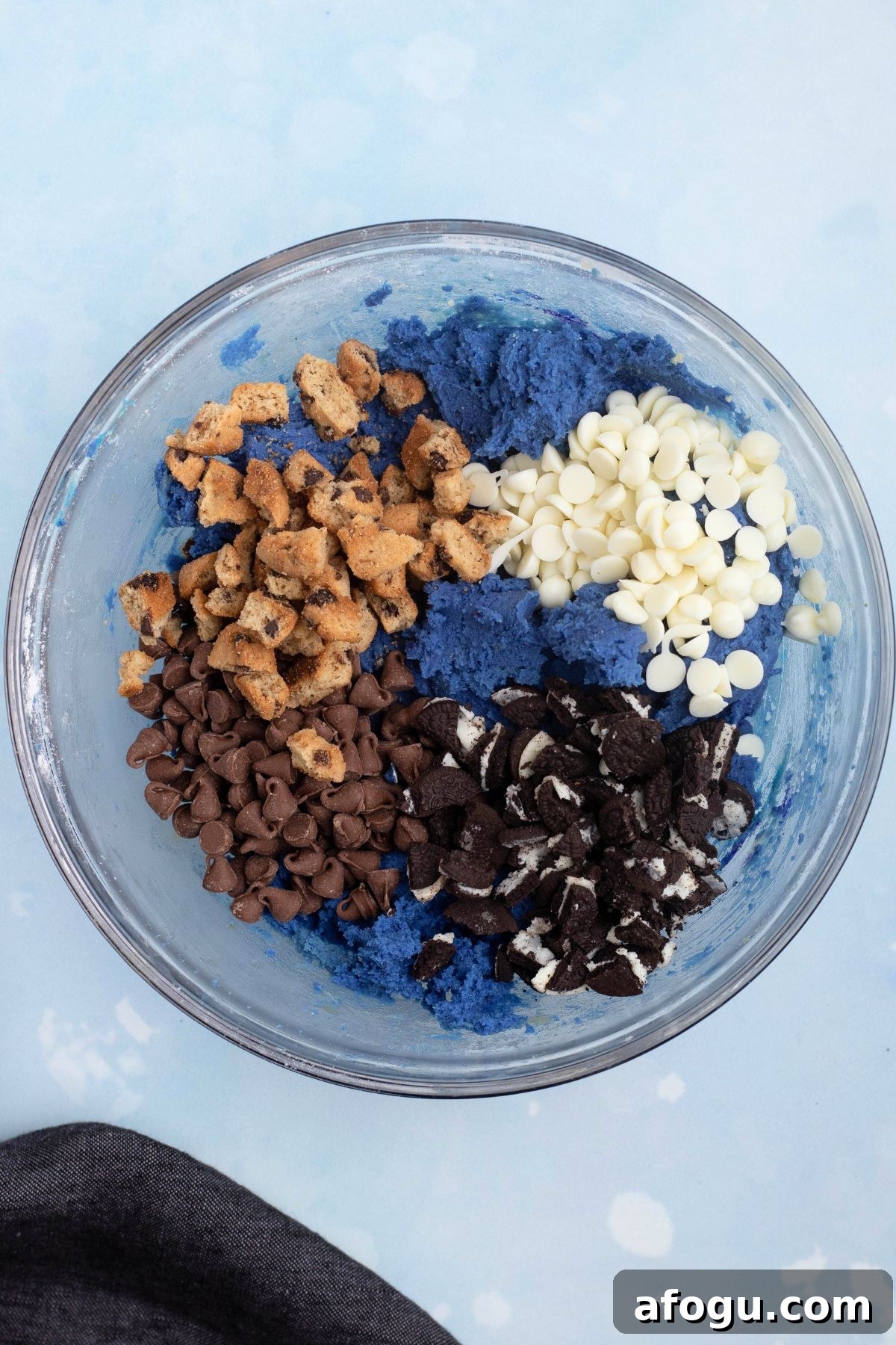 Chopped cookies and chips being mixed in with cookie dough in a clear glass bowl.