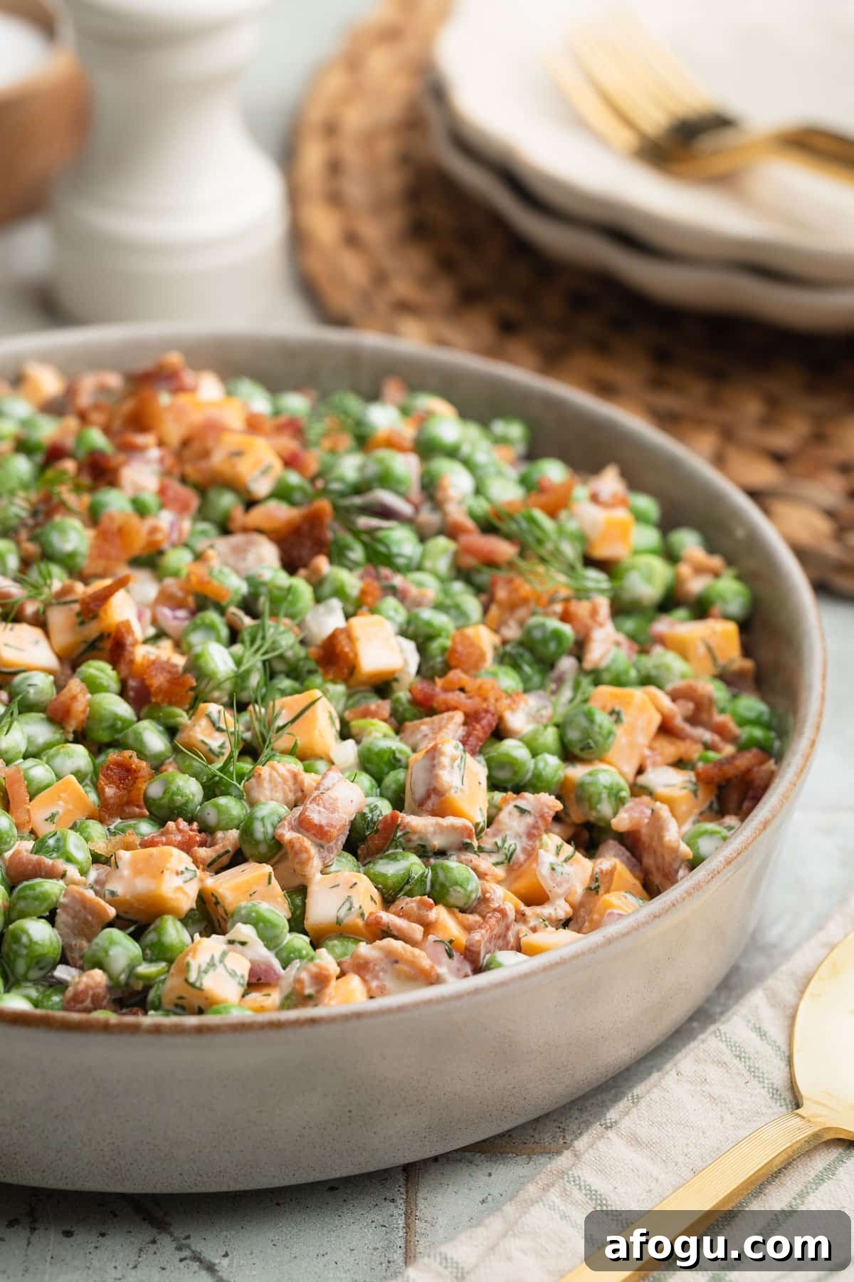 Close-up photo of creamy pea salad served in a shallow ceramic bowl, made with green peas, cheddar cheese cubes, crispy bacon, chopped pecans, diced red onion, and fresh dill tossed in a creamy dressing. Gold flatware and stacked plates sit blurred in the background.